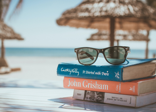 A stack of colorful books and sunglasses on a table, with a beach view in the background.